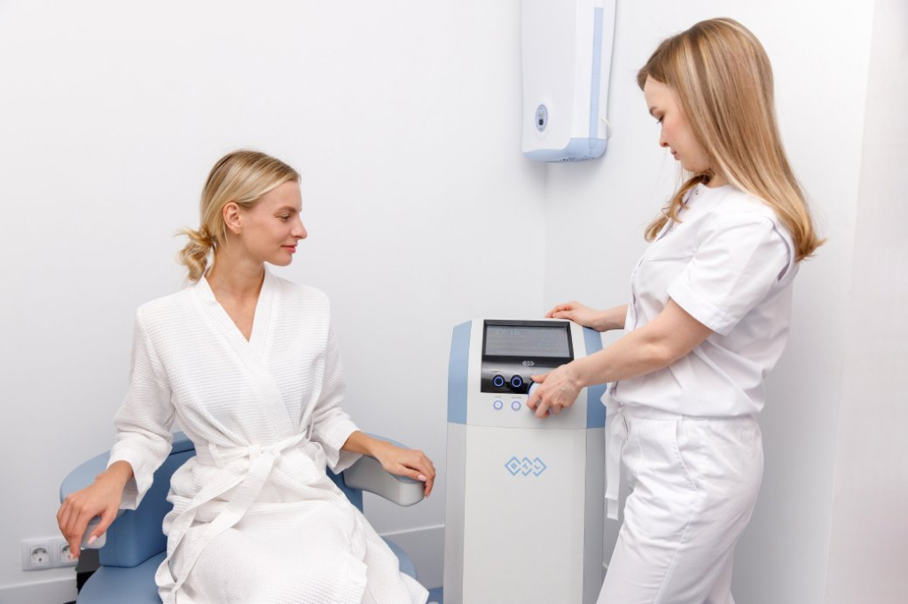 A medical professional operates a piece of medical equipment while a patient sits nearby.